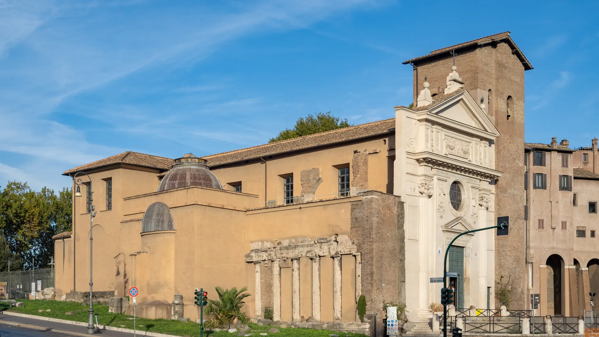 Basilikaen San Nicola in Carcere i Rom i aftenlys: en lys barokfacade, et middelalderligt murstensklokketårn og, ved siden af kirken, en række antikke tempelsøjler indbygget i dens mur.